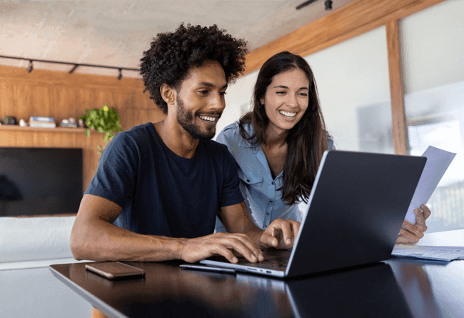 Duas pessoas juntas em frente a um laptop em um ambiente doméstico com decoração moderna e plantas ao fundo.